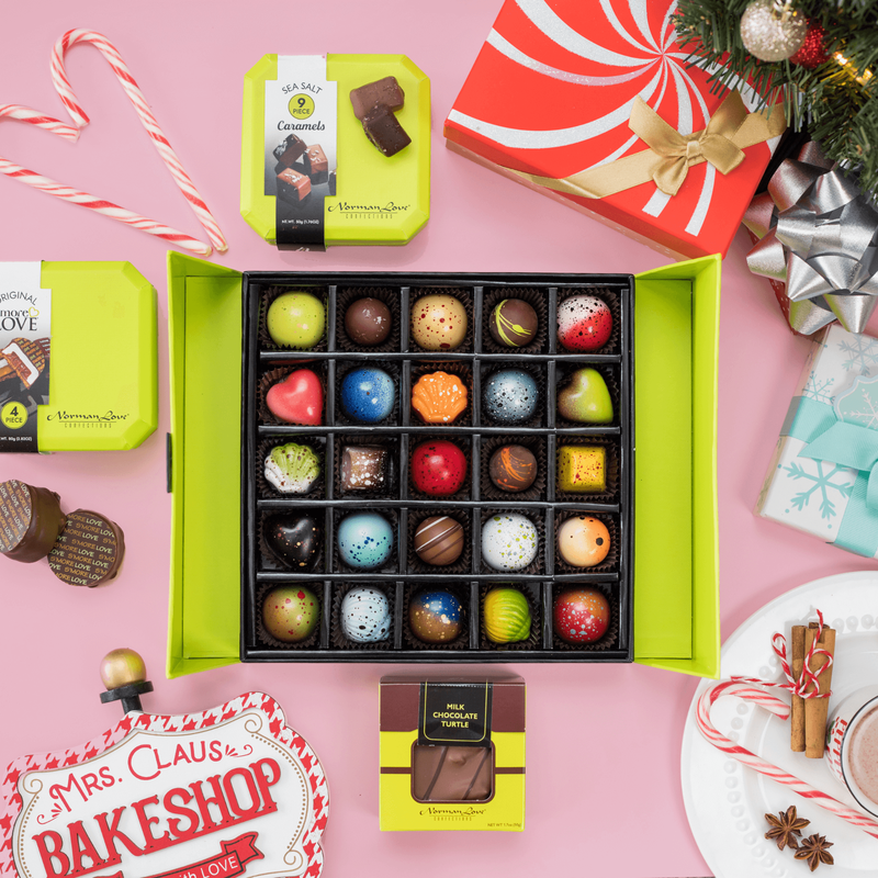 A close-up an open 25 piece holiday gift box on a pink background. The gift box displays beautifully crafted chocolates including all 10 Christmas inspired flavors. Christmas decorations and the contents of the Classic Treats Bundle surround the box.